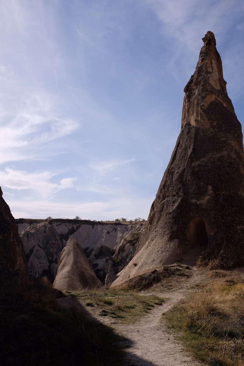 A tall, conical rock formation dominates the right side of the frame, with a dirt path winding through the foreground under a light blue sky.