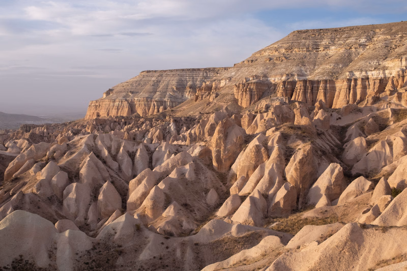A panoramic view of dramatic badlands featuring numerous conical hills and layered sedimentary rock formations under a pale sky.
