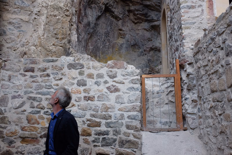An older man looks up at a stone wall with a narrow arched doorway in an old stone building.