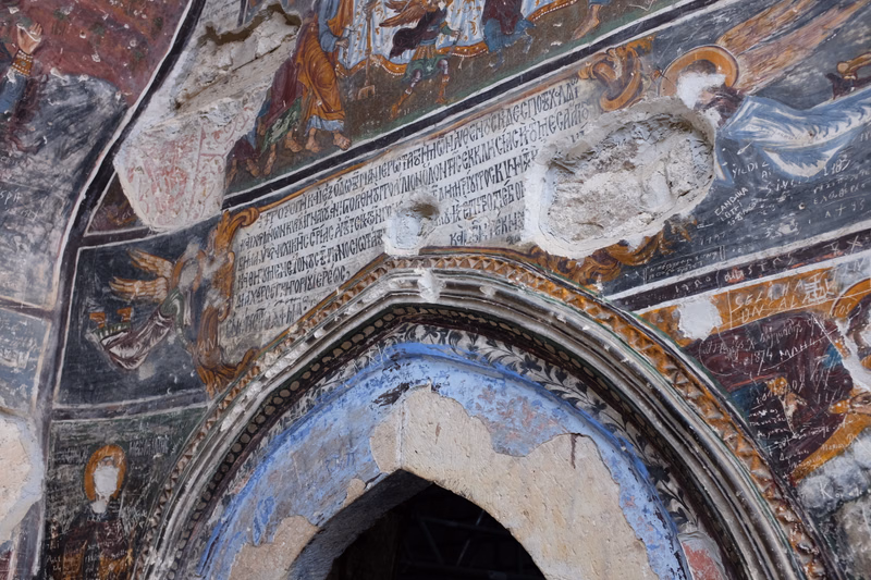 An interior view of a church featuring faded frescoes covering the walls, with a prominent arched doorway below.