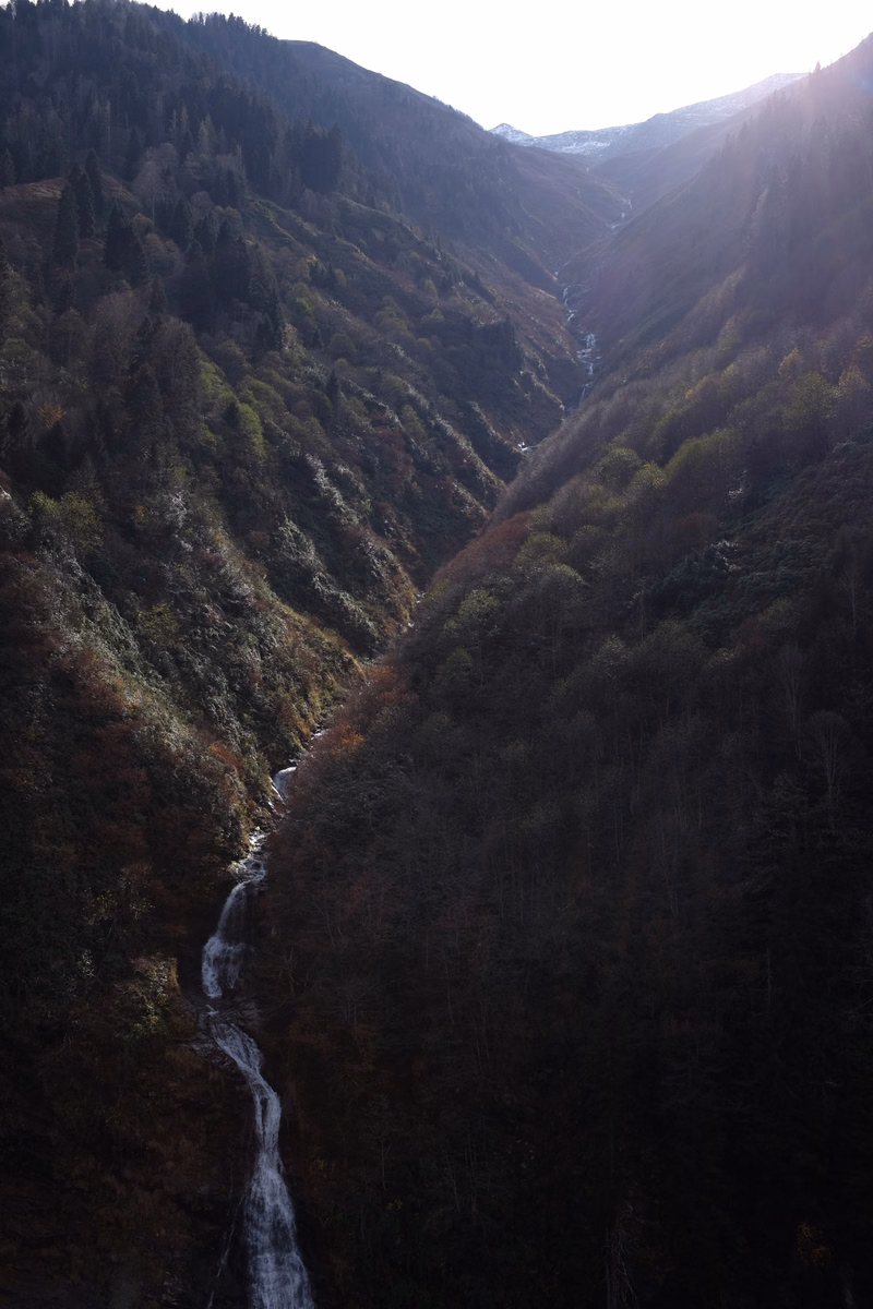 A narrow, steep valley with dense forest slopes features a cascading waterfall flowing down the mountainside under bright sunlight.
