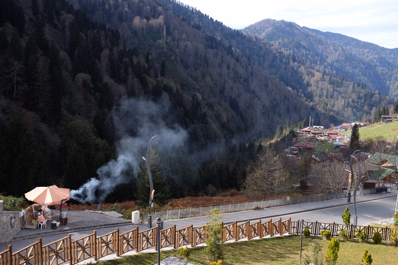 Smoke rises from an outdoor restaurant area situated near a road with a backdrop of forested mountains.