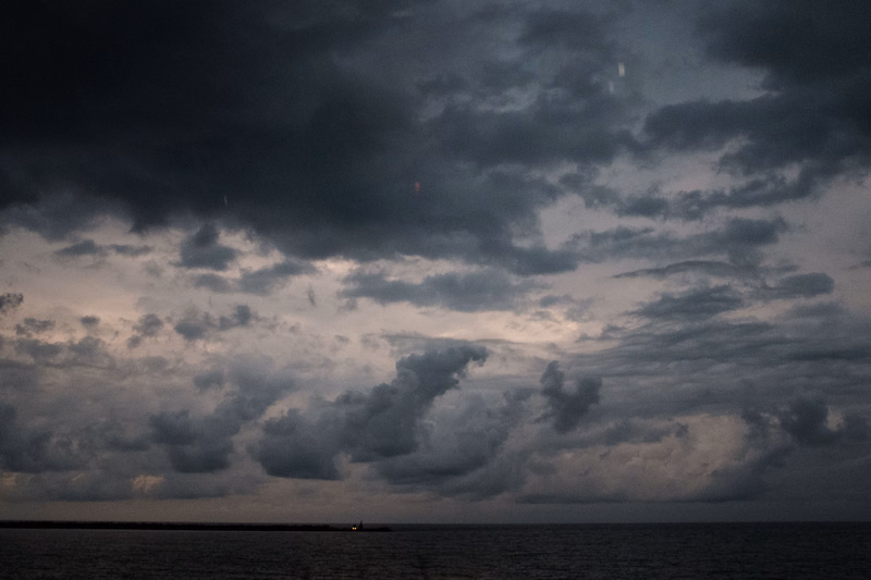A dark, dramatic sky filled with heavy clouds hangs over a calm ocean with a small lighthouse visible on the horizon.