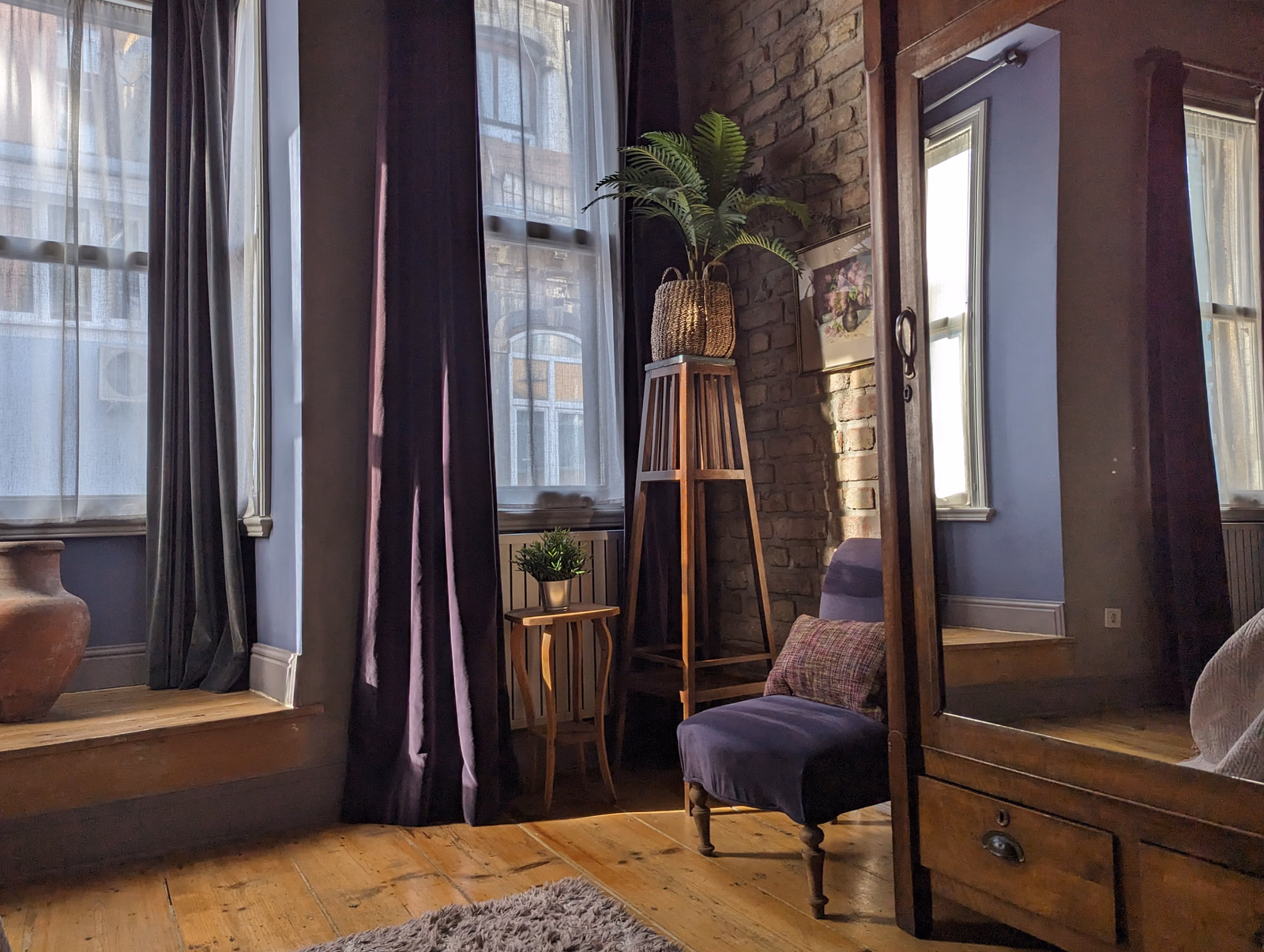 A room with large windows draped in dark curtains, featuring a wooden mirror and a small upholstered chair.