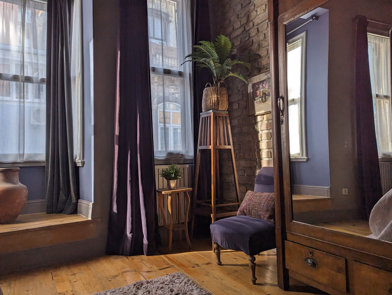 A room with large windows draped in dark curtains, featuring a wooden mirror and a small upholstered chair.