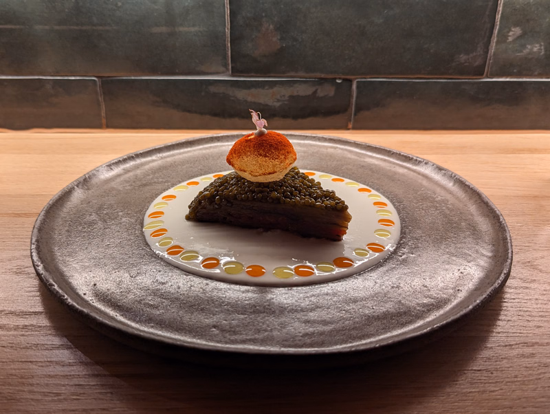 A slice of dark cake topped with a small round pastry sits on a decorative white plate, which is placed on a gray textured plate.