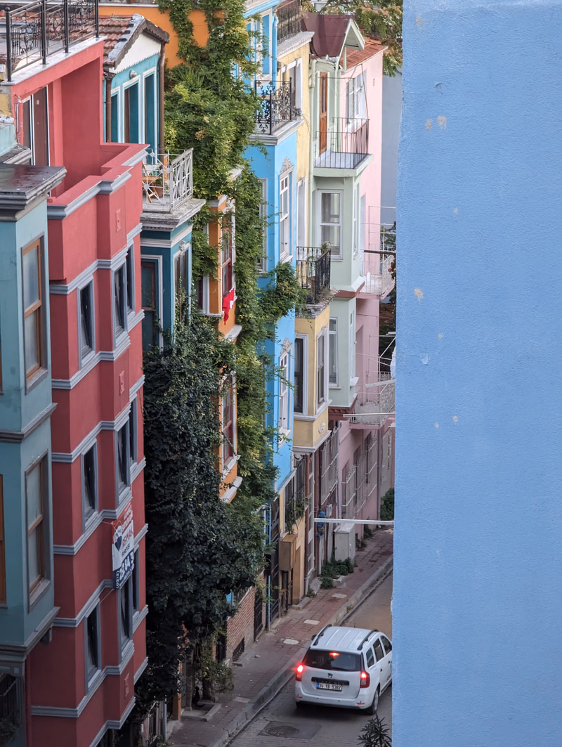 A row of colorful, multi-story apartment buildings lines a narrow city street, with a white car parked near the curb.