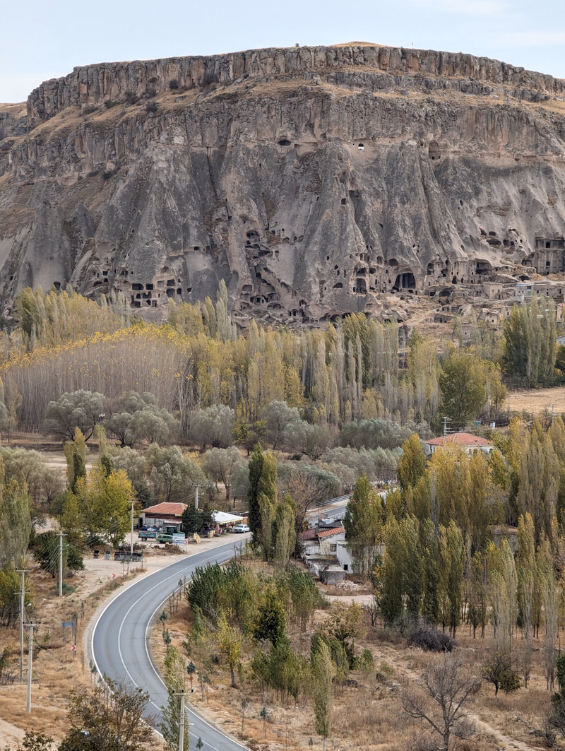 A large, rocky cliff face with numerous cave dwellings overlooks a valley containing a winding road and scattered trees.