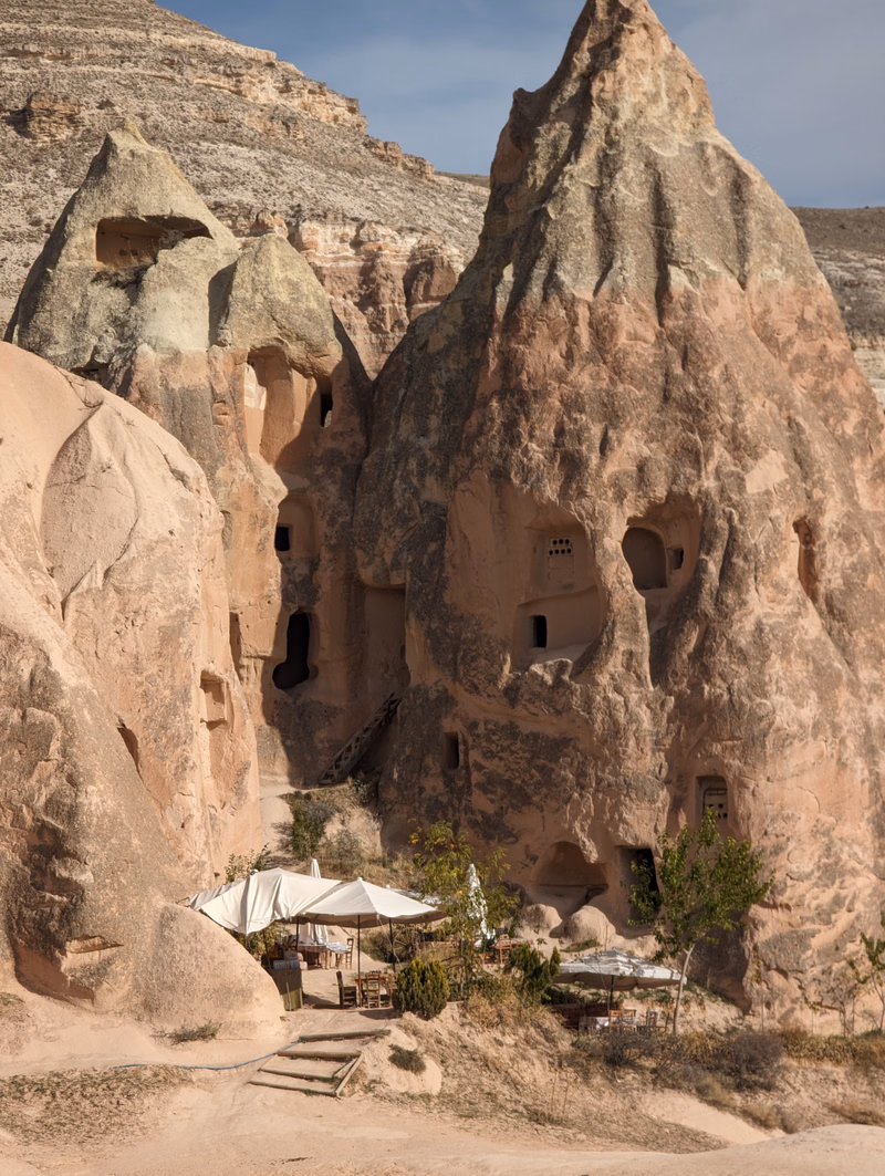 A cluster of ancient cave dwellings carved into large, reddish-brown rock formations, with small tents set up at the base.