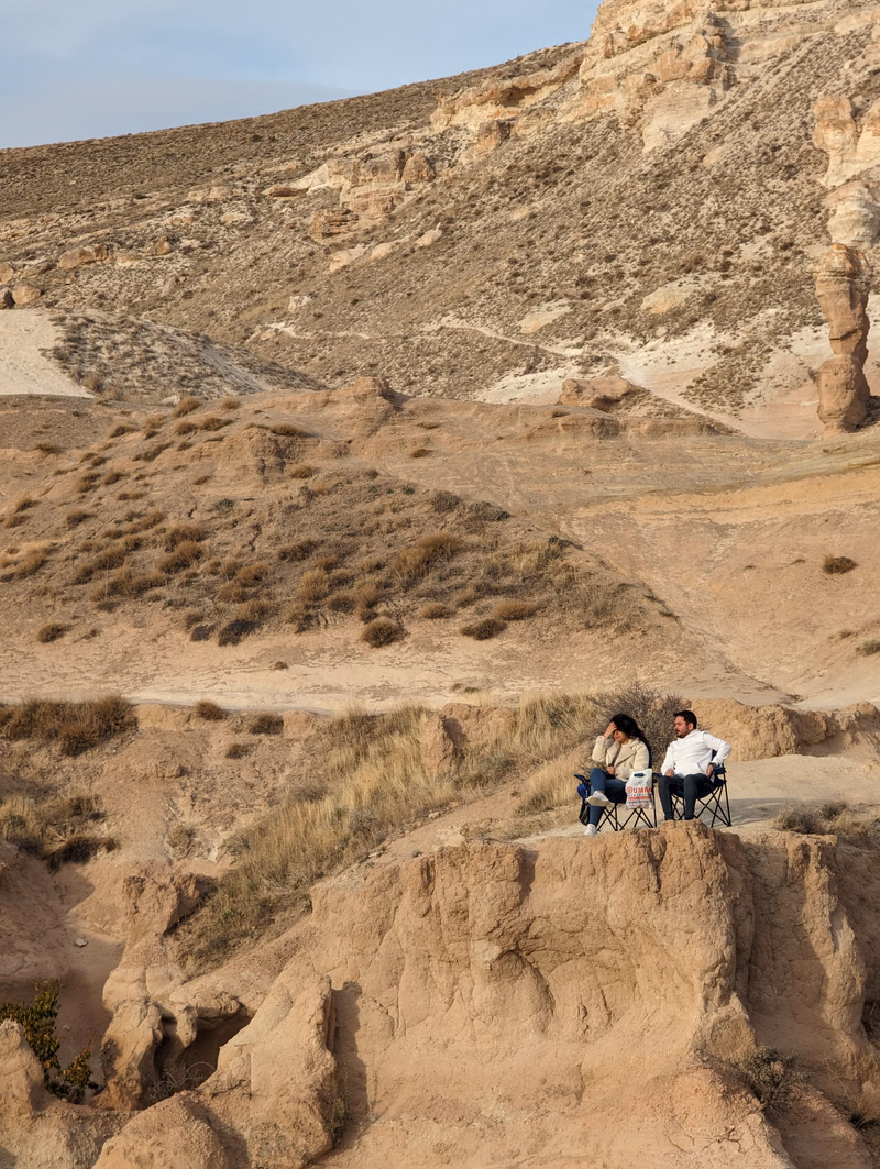 Two people sit on a sandy cliff edge overlooking a dry, hilly desert landscape.