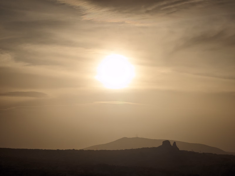 A bright sun shines over a hazy desert landscape with silhouetted hills and distant mountains.