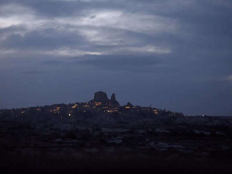 A silhouette of a city nestled among hills under a dramatic, cloudy night sky with scattered lights visible.