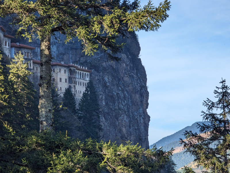 A medieval castle is built into the side of a massive, sheer rock cliff, framed by evergreen trees under a blue sky.