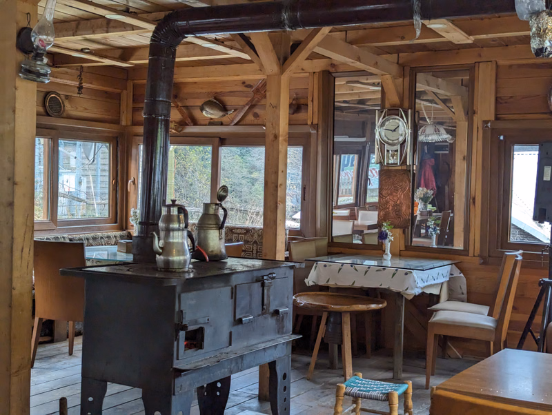 A large, black wood stove with copper kettles sits in a rustic wooden cabin kitchen, surrounded by wooden beams and windows.