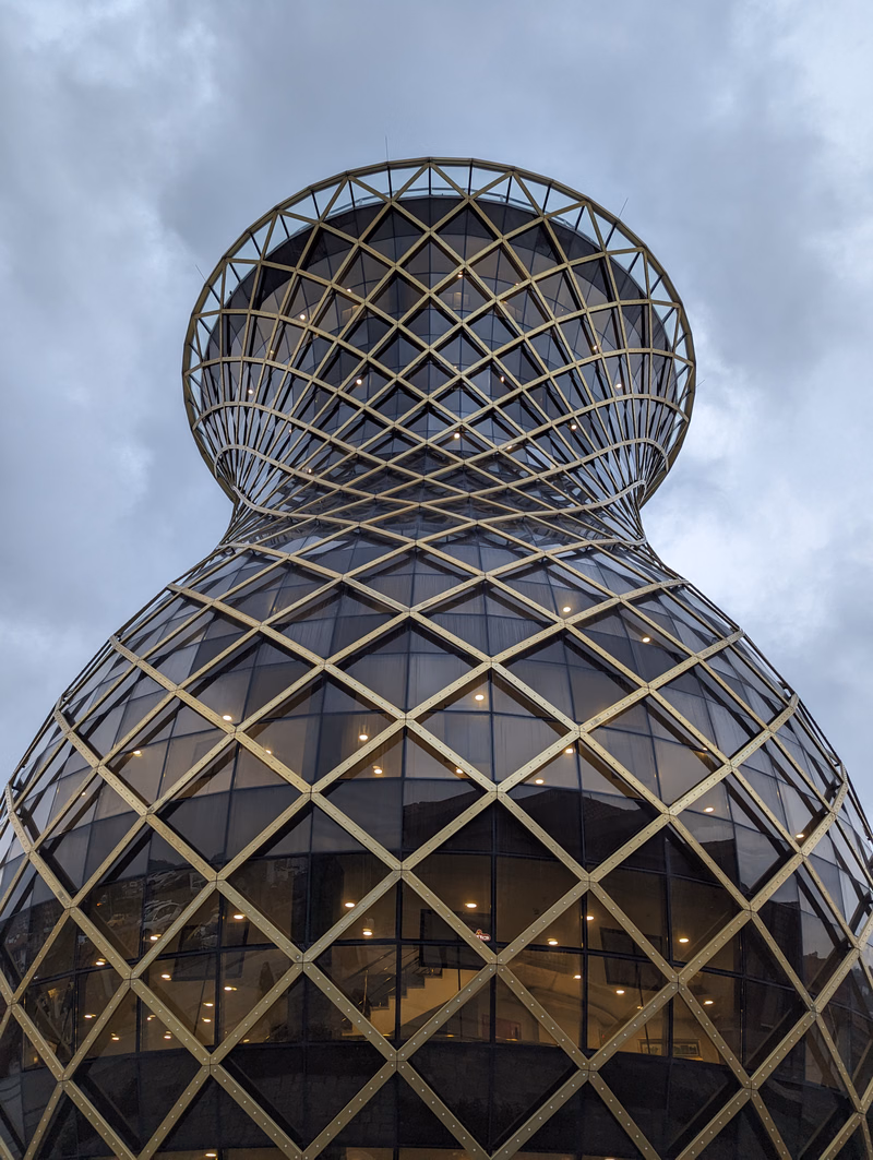A large, modern glass dome building with a complex geometric, latticed exterior is viewed against a cloudy sky.