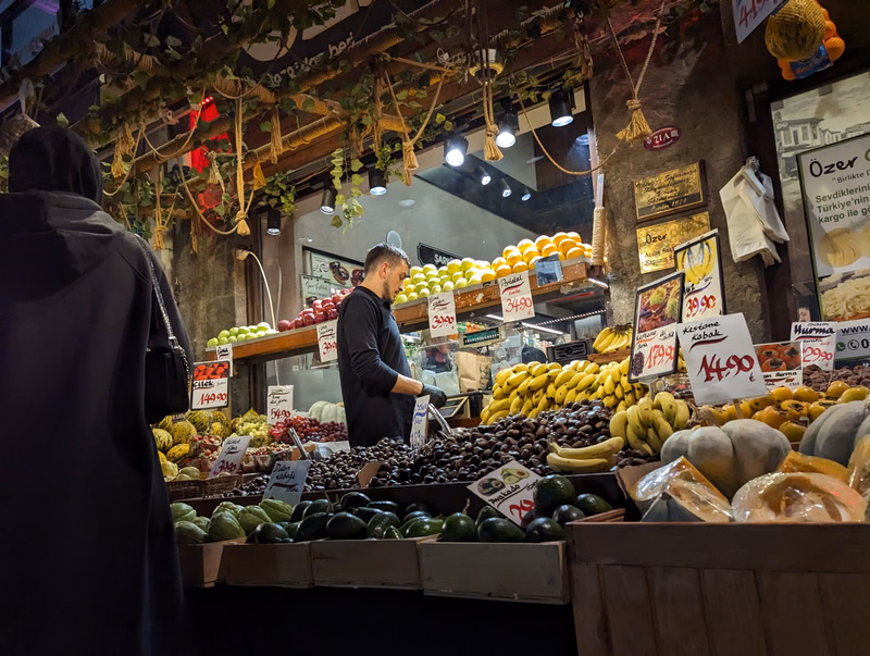 A male vendor in a market stall displays various fresh fruits and vegetables, including bananas and avocados, with signs like "14.90" visible.