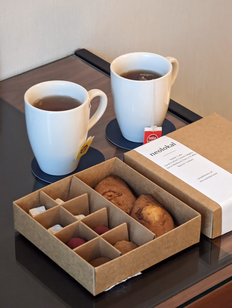 A set of white mugs containing tea is displayed next to a cardboard box filled with cookies, with a small card reading "neolokal" visible.