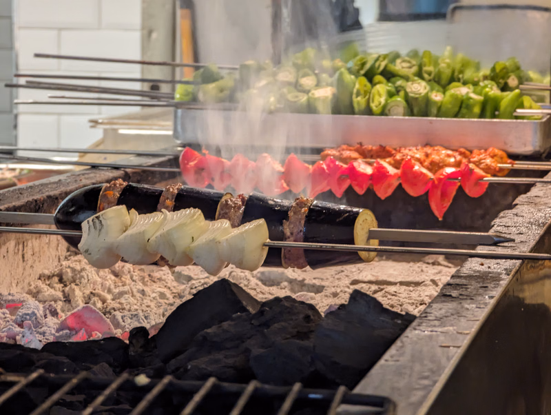 Various skewers of meat and vegetables are cooking over hot coals in a metal grill setup.