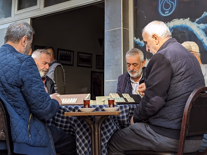 Several older men are gathered around a small table in a cafe, playing a card game.