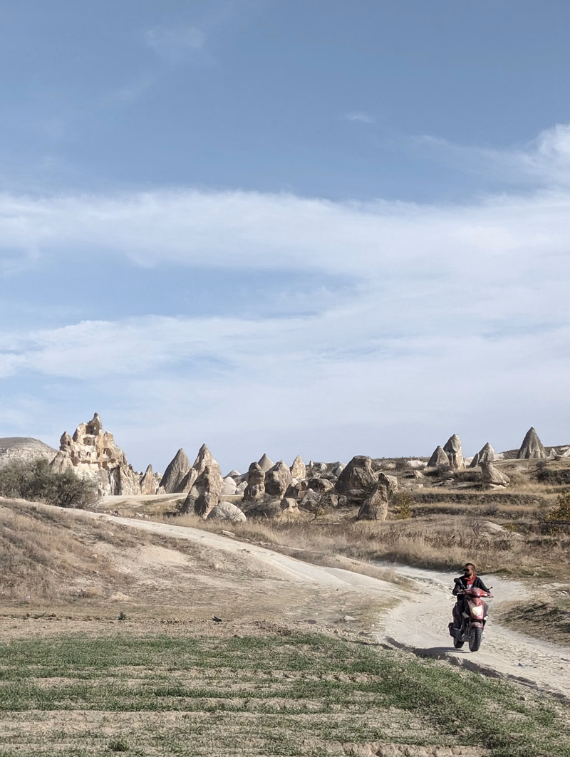 A motorcyclist rides on a dirt road through a landscape featuring numerous conical rock formations under a bright sky.