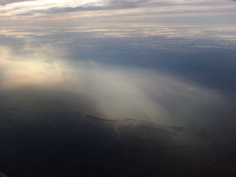 A dense layer of fog hangs over a coastline as the sun rises, casting beams through the mist.