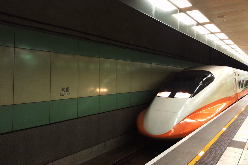 A modern high-speed train with orange accents is stopped at a subway platform, with a sign reading "桃園 Taoyuan" on the wall.