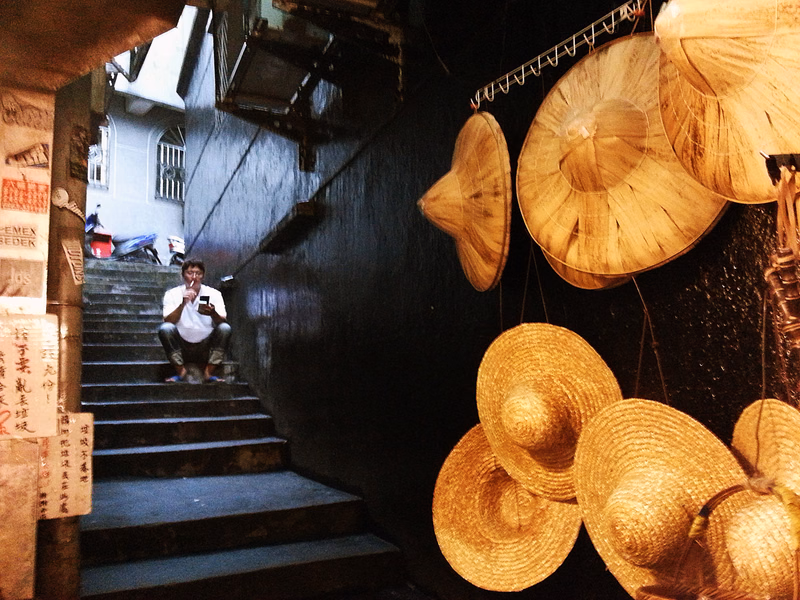 A man sits on stone stairs in an alleyway next to a display of various straw hats hanging against a dark wall.