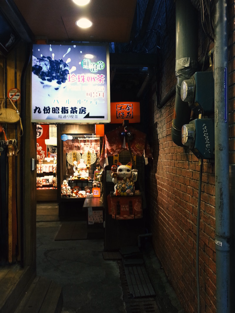 A Japanese tea shop with a sign that reads "九份茗茶" is visible in a narrow alleyway next to a brick wall.