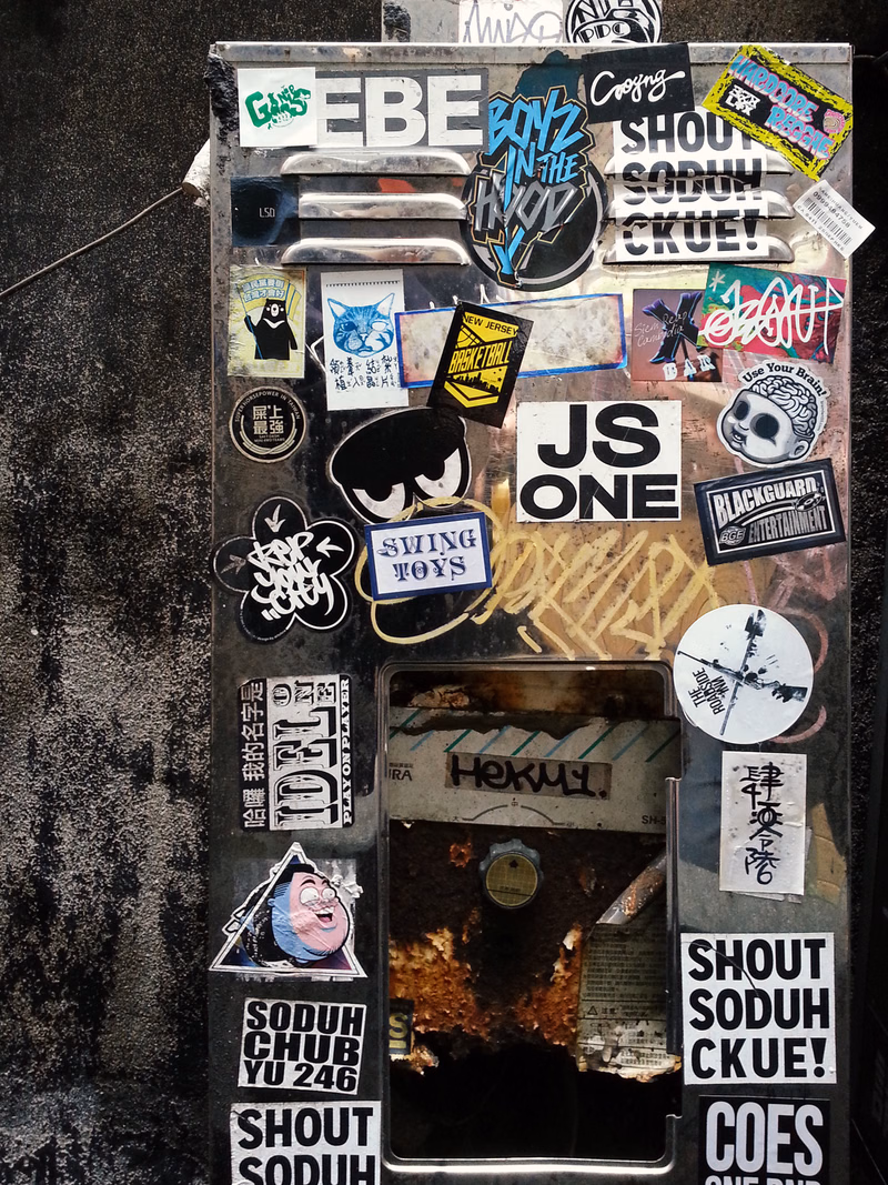 A collection of stickers and graffiti covers a metal box, featuring text like "EBE BONZ IN THE SKY" and "JS ONE".