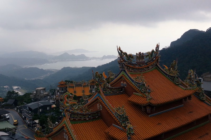A large, ornate temple with a tiled roof overlooks a misty bay surrounded by green mountains under a cloudy sky.