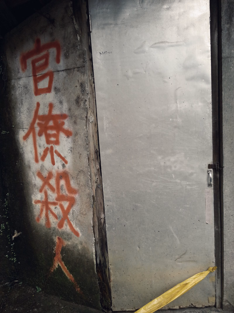 A weathered metal door has red graffiti written vertically on its left side, which reads "官係殺".