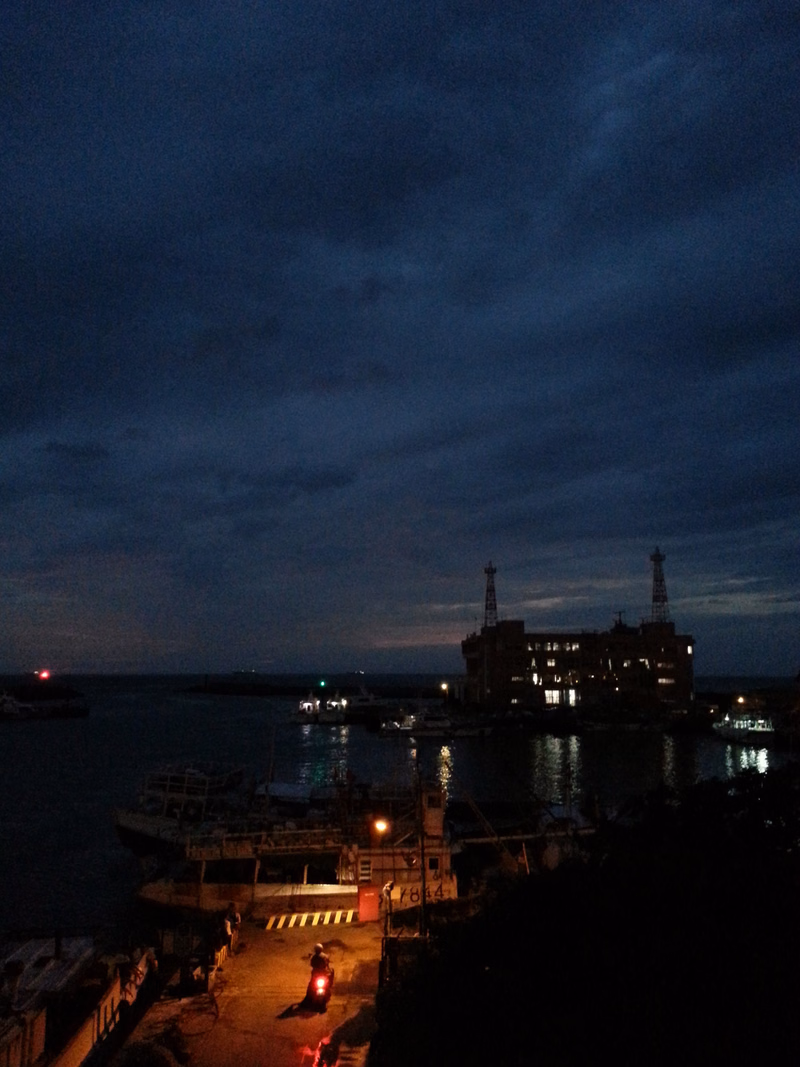 A nighttime view of a harbor with industrial buildings illuminated against a dark, cloudy sky.