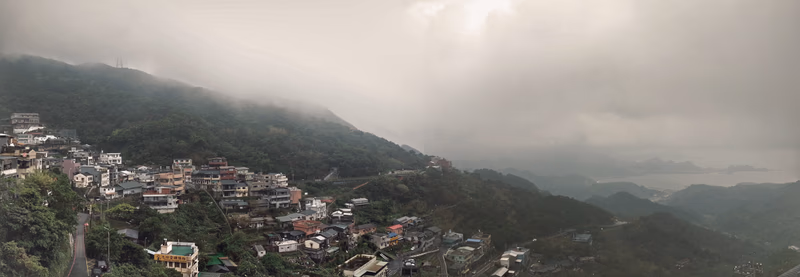 A panoramic view of a village nestled in the mountains with a cloudy sky and a road winding through the village.