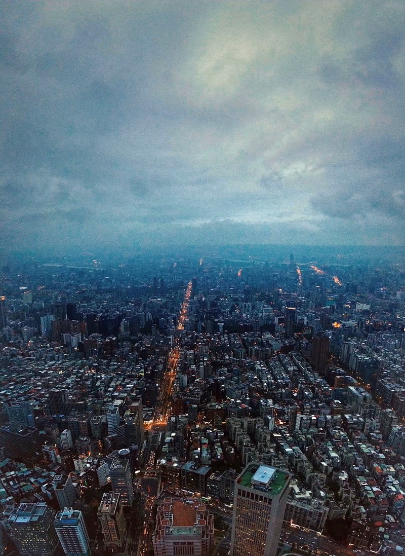 A high-angle view of a sprawling city at night under a heavily clouded, dark sky, with streaks of light visible in the distance.