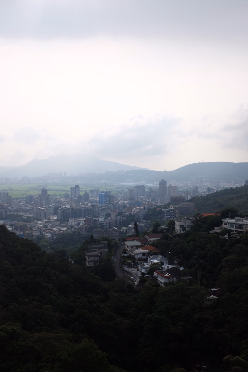 A hazy cityscape stretches across a valley, with dense urban development nestled among rolling, forested mountains.