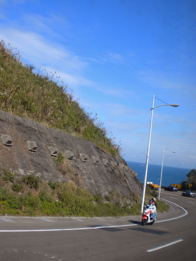 A motorcycle travels along a winding coastal road with a steep, vegetated hillside and blue sky overhead.