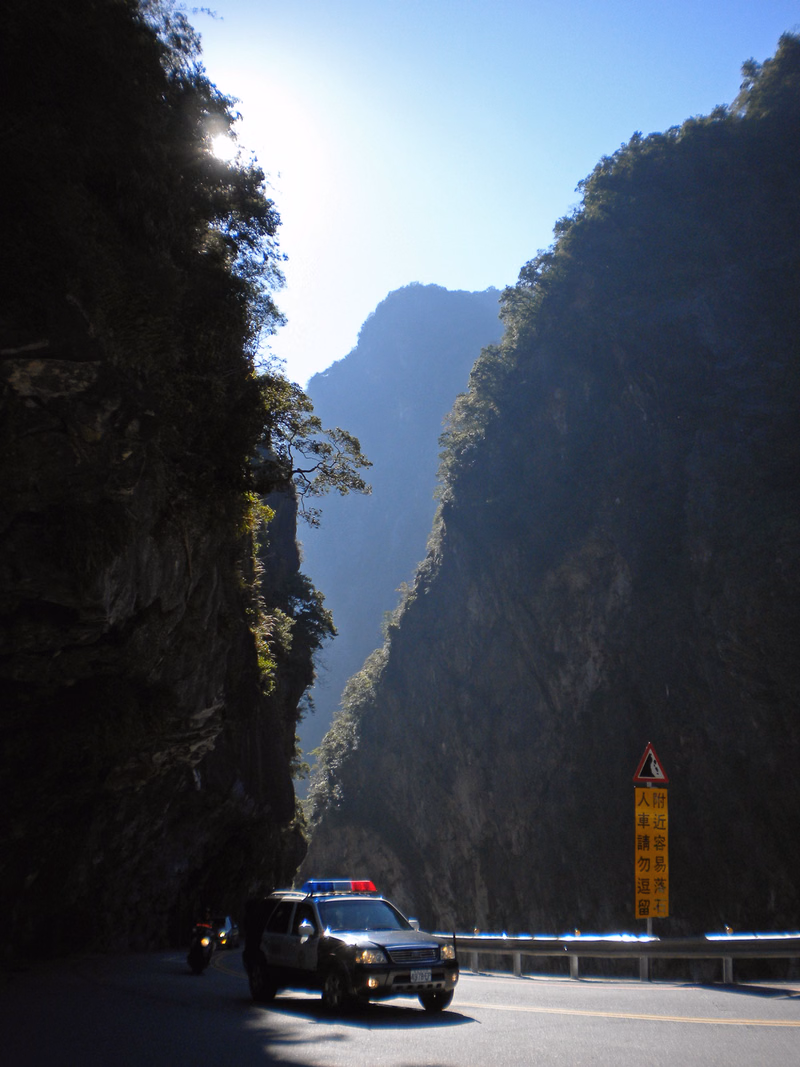 A car drives along a winding mountain road flanked by steep, forested cliffs under a bright sky.