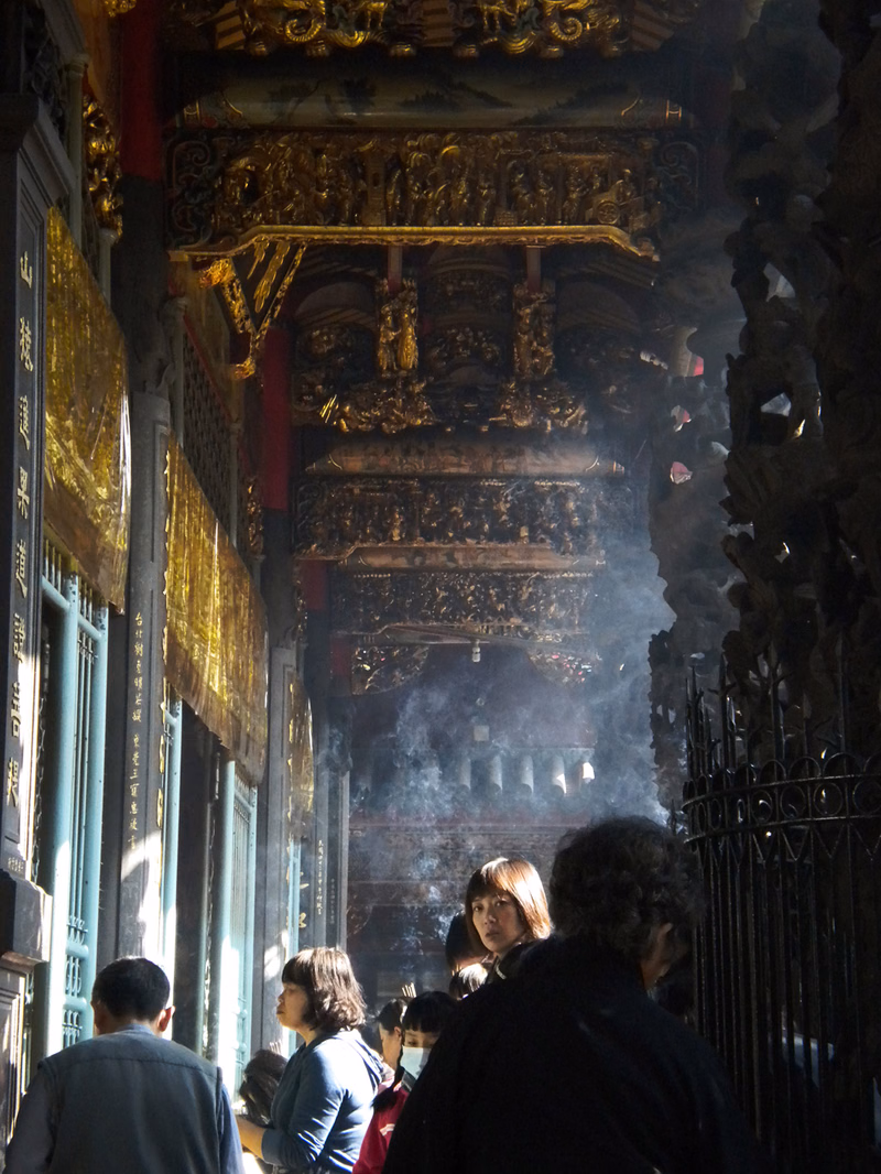 A view inside a richly decorated temple featuring a large, ornate golden statue under a high ceiling, with visitors gathered below.