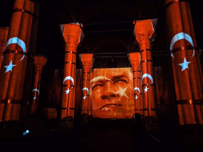 A large projection of a historical figure's face is displayed on a wall in the Eminönü district of Istanbul, Turkey. The columns surrounding the space are adorned with the national flag of Turkey.