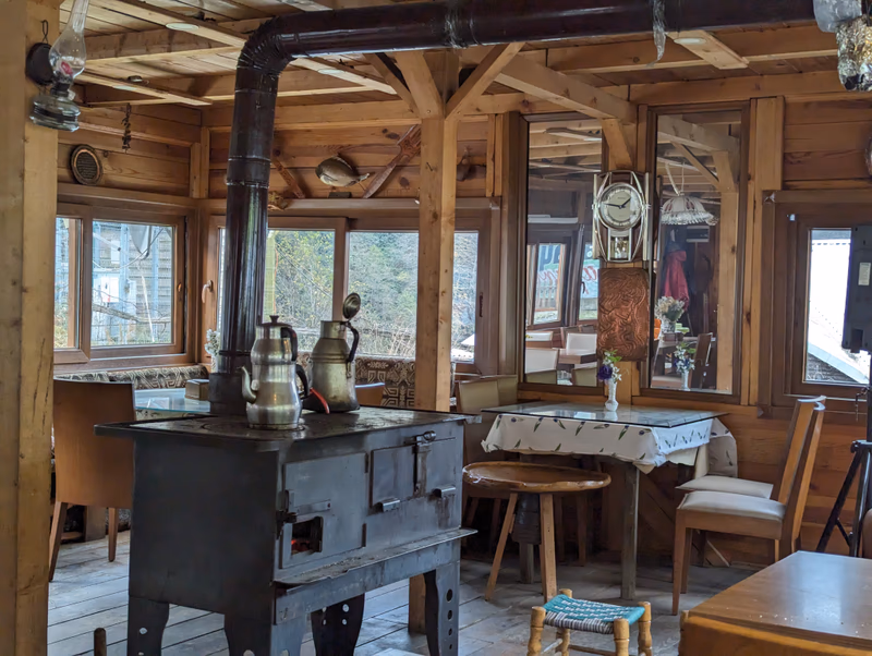 A rustic, wooden interior with a wood-burning stove in the foreground. The room is filled with various objects, including a kettle, a table with a tablecloth, and a few chairs. The walls are adorned with artwork and a clock, and the windows provide a view of the outdoors.
