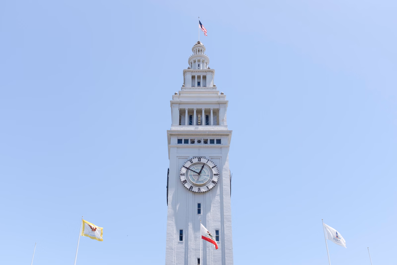 A tall, white city hall with a prominent clock tower stands against a clear, light blue sky, flying an American flag.