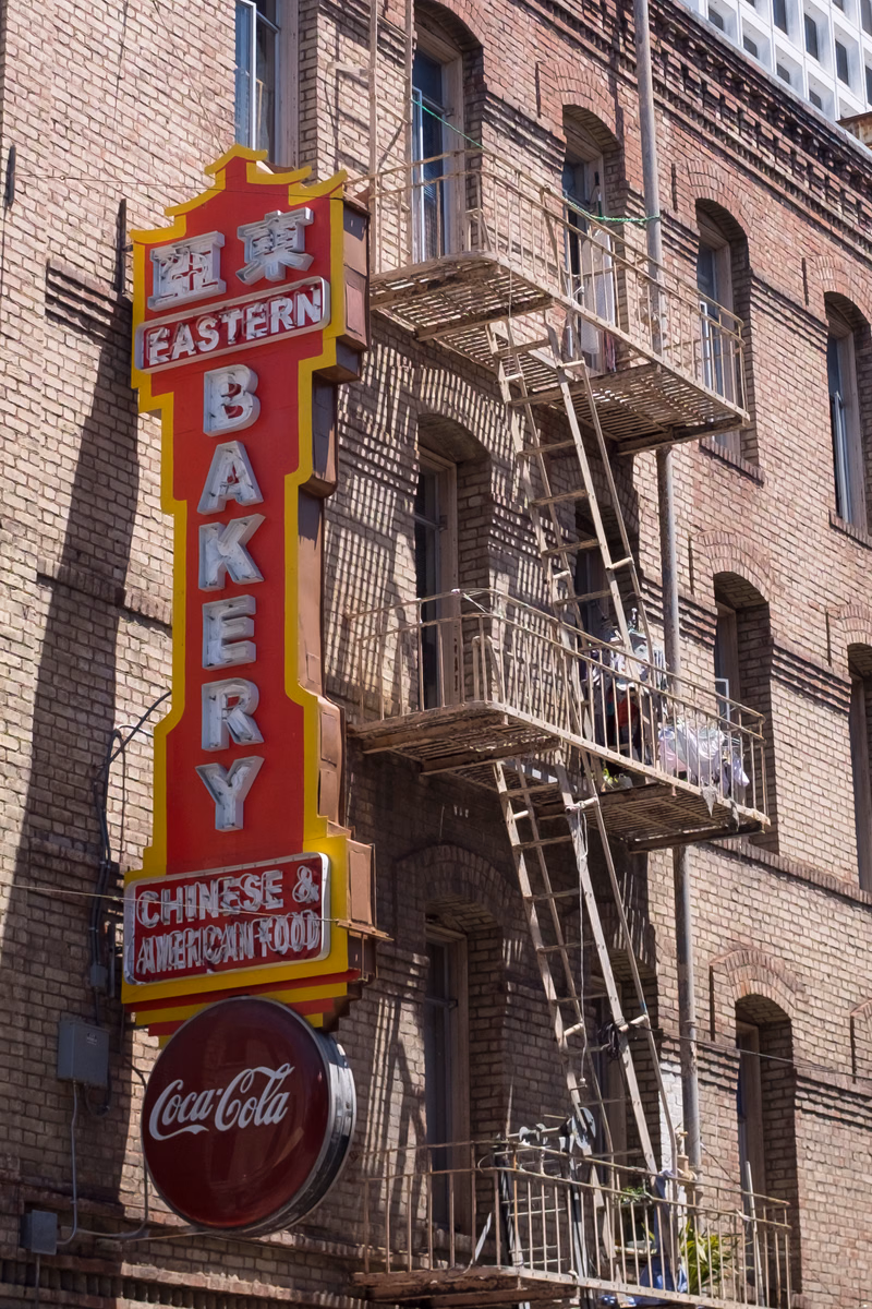 A large vertical sign for "EASTERN BAKERY" with "CHINESE & AMERICAN FOOD" below, next to a brick building with an external metal staircase.