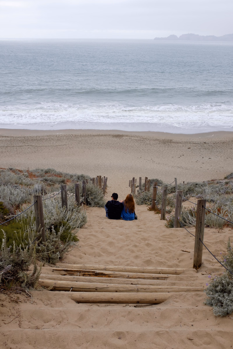 A couple sits on the sand near the ocean, with wooden planks laid out in front of them.