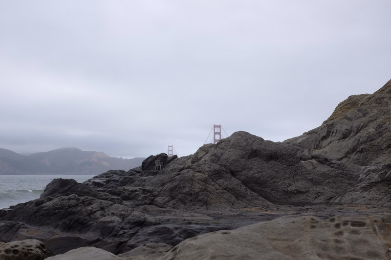 The Golden Gate Bridge is visible through heavy fog above dark, rocky coastal terrain.