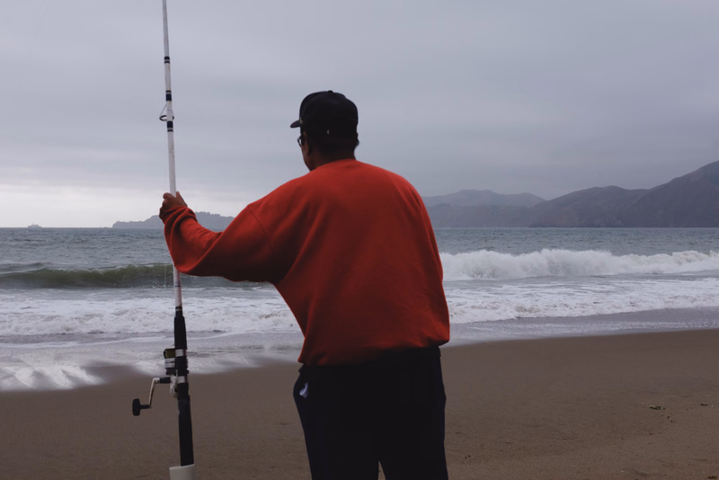 A man wearing a red sweater stands on a sandy beach holding a fishing rod while looking out at the ocean under a cloudy sky.