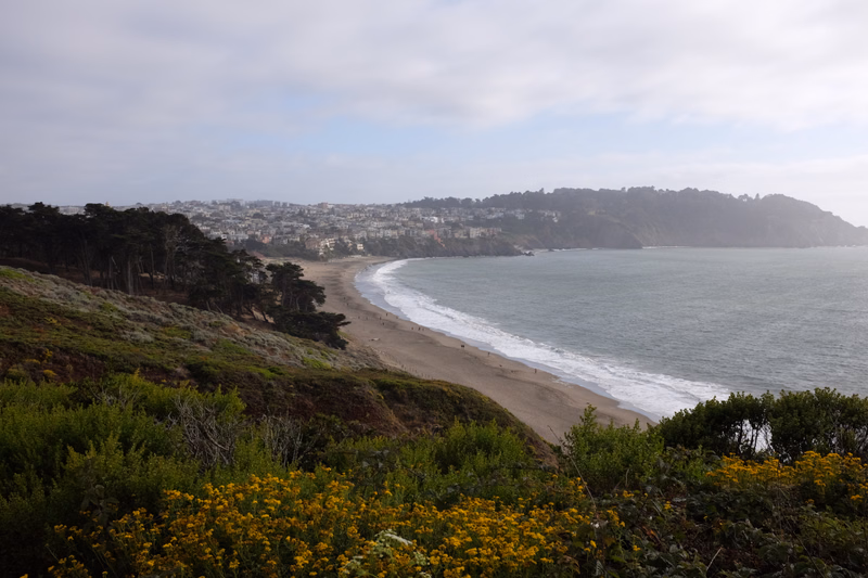 A wide view of a sandy beach curving along a coastline with a city built into the hills under a cloudy sky.