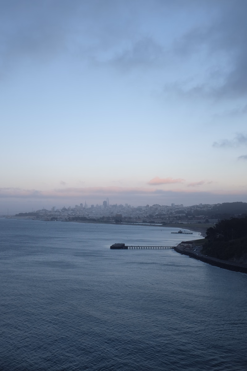 A wide view of a coastal city skyline under a pale, cloudy sky, featuring a long pier extending into the water.