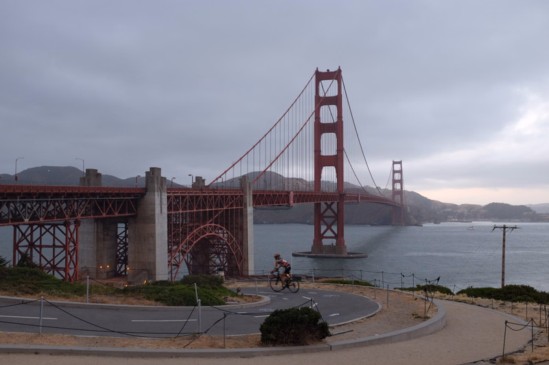 A cyclist rides near the base of the iconic red Golden Gate Bridge under an overcast sky.