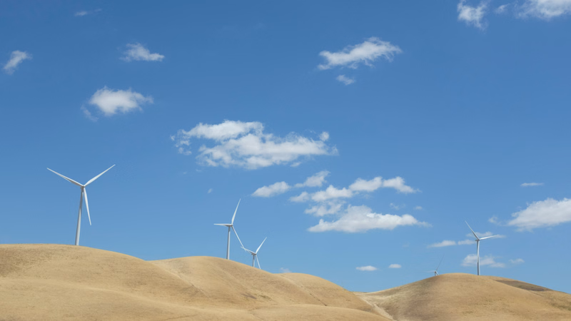 Several modern wind turbines are visible atop rolling, sandy hills under a bright blue sky with scattered white clouds.