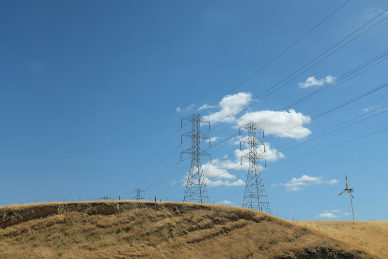 High-voltage power lines stretch across a grassy hill under a bright blue sky, with a wind turbine visible on the right.